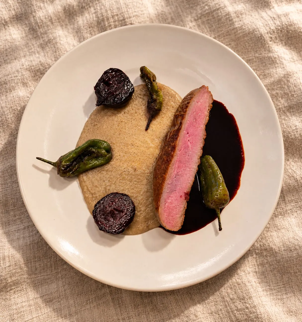 Une tranche de viande rosée sur une purée crème, avec des betteraves rôties, des poivrons verts et une sauce sombre, dans une assiette crème posée sur du lin
